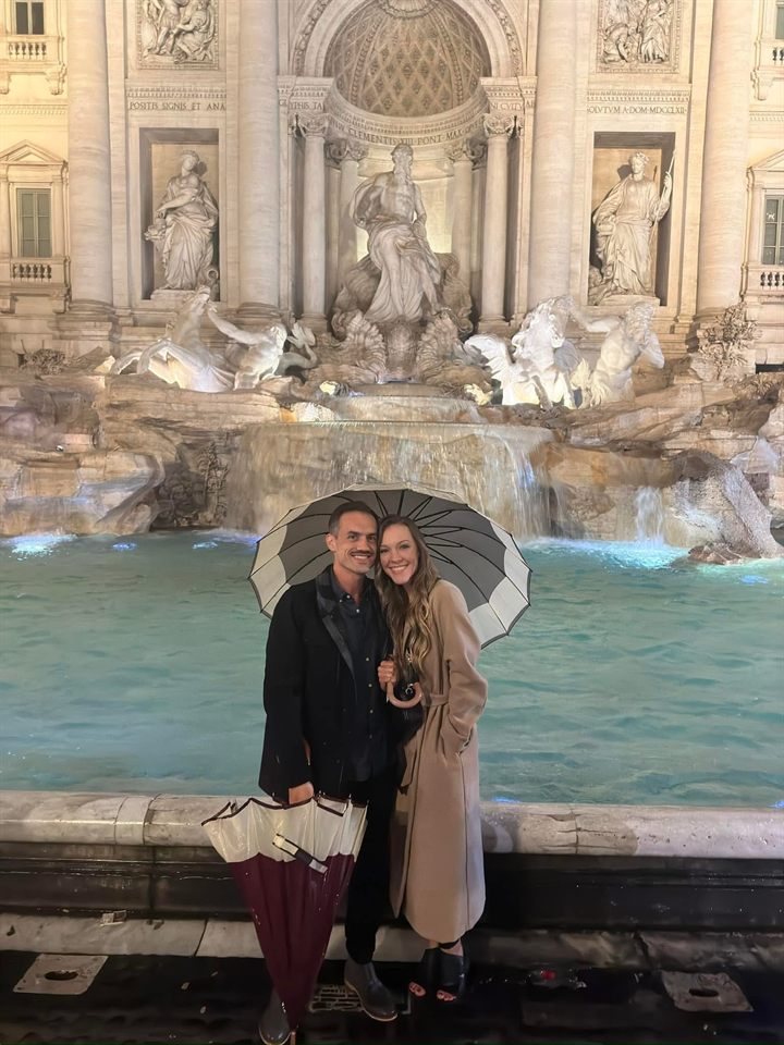 Taylor and his wife sharing an umbrella in front of the Trevi Fountain at night.